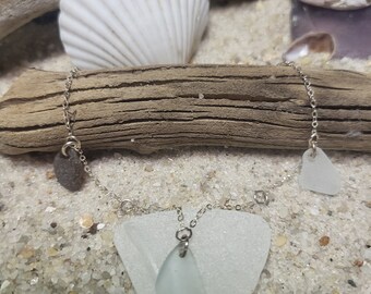 May include: A silver chain necklace with three sea glass pendants. The pendants are a teardrop shape, a small triangle, and a larger triangle. The necklace is displayed on a piece of driftwood with seashells.