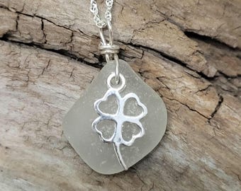 May include: A silver-toned necklace featuring a four-leaf clover pendant. The pendant is a silver-colored charm attached to a piece of sea glass. The necklace chain is also silver-colored. The background is a wooden surface.