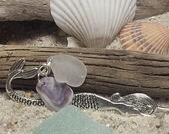 May include: A silver mermaid bookmark with a purple and white seashell charm and two pieces of sea glass. The bookmark is lying on a bed of white sand.