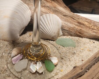 May include: A set of six gold-colored wine glass charms featuring seashells and sea glass. The charms are hanging from a wine glass.