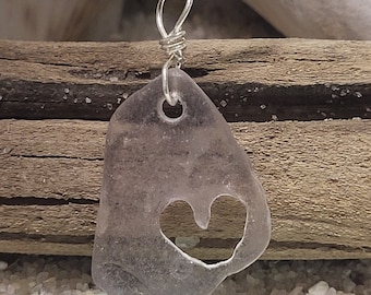 May include: A clear glass pendant with a heart-shaped cutout, hanging from a silver chain. The pendant is resting on a green piece of sea glass.