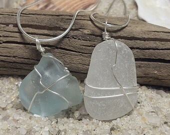 May include: Two sea glass pendants on silver chains. One pendant is a light blue, the other is clear. Both are wire-wrapped and rest on a piece of driftwood, surrounded by sand and seashells.