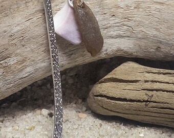 May include: Silver bookmark with a decorative design and a charm featuring a pink and brown seashell.