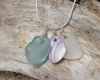 May include: A silver chain necklace with three charms: a light blue sea glass charm, a purple and white seashell charm, and a white sea glass charm.
