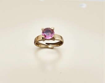 May include: A gold-toned ring featuring a large, round, pink gemstone. The ring's band has a simple, elegant design with a split shank. The gemstone is held in place by four prongs. This ring is a piece of jewelry.