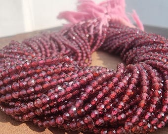 May include: A strand of deep red faceted gemstone beads. The beads are small and round, and they are strung together on a clear thread. The beads are arranged in a circular pattern, and they are all of the same size and shape.