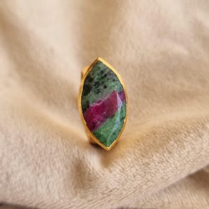 Puede incluir: Un anillo con forma de marquesa que presenta una piedra preciosa facetada de rubí y zoisita engastada en una banda de metal dorado. La piedra muestra un patrón de color distintivo con secciones magenta intenso frente a inclusiones verde oscuro.
