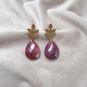 Op de afbeelding: Een paar goudkleurige oorbellen in de vorm van bladeren met gefacetteerde, druppelvormige, rode edelstenen. De oorbellen hangen aan een goudkleurig bladerontwerp. De edelstenen hebben een dieprode kleur en zijn geslepen om het licht te vangen.