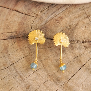 Könnte beinhalten: Ein Paar goldfarbene Ohrringe mit einem fächerförmigen Oberteil, das einen kleinen, klaren Stein enthält, verbunden mit einem dünnen Stiel, der in einer hängenden, runden, hellblauen Perle endet. Das Design ähnelt einer stilisierten Pflanze.