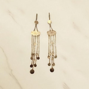 May include: A pair of gold-coloured earrings. Each earring has a small circular top, a textured oval-shaped centrepiece, and multiple dangling chains with small, round, flat charms. The earrings are set against a white marble background.
