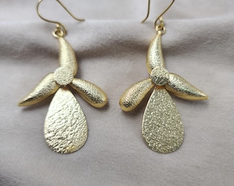 May include: A pair of gold-colored earrings. Each earring features a textured, teardrop-shaped pendant suspended from a floral design with three petals. The earrings have a hook closure.