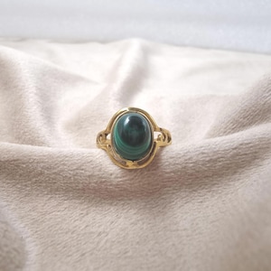 May include: A gold ring with a dark green malachite stone. The oval stone is set in a decorative, openwork gold band. The ring is presented on a soft, neutral-toned fabric.
