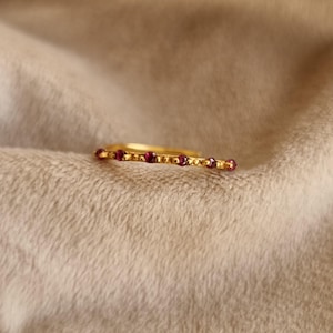 May include: A gold-colored ring with a row of small, round, red gemstones. The ring is set against a soft, neutral-toned background, highlighting the jewelry's delicate design and color.
