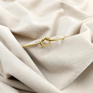May include: A gold-coloured bracelet featuring a knot design. The bracelet is crafted from a thin, smooth metal and is displayed against a soft, cream-coloured fabric. The knot is the central feature of the bracelet.