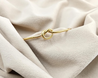 Peut inclure: Un bracelet doré avec un motif de nœud. Le bracelet est fait d'un métal fin et lisse et est posé sur un tissu doux de couleur crème. Le nœud est le point central du bracelet.
