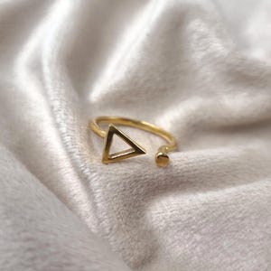 Puede incluir: Un anillo dorado con un diseño abierto. El anillo presenta una forma triangular en la parte delantera y un pequeño detalle redondo en el extremo abierto. El anillo está sobre un fondo blanco suave.
