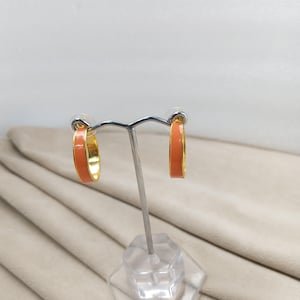 May include: A pair of gold-tone hoop earrings with a vibrant orange enamel finish. The earrings are displayed on a silver earring stand against a neutral background. The hoops have a classic design, showcasing a pop of color.