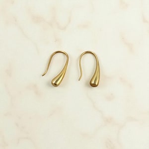 Peut inclure: Une paire de boucles d'oreilles dorées en forme de goutte d'eau. Les boucles d'oreilles ont un design en forme de crochet incurvé et une finition lisse et polie. La forme de goutte d'eau est allongée, avec un fond arrondi. Elles sont posées sur une surface claire, marbrée.