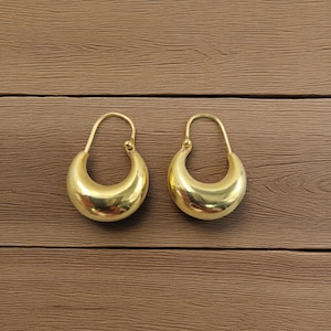 May include: A pair of gold-coloured, crescent-shaped hoop earrings. Each earring features a rounded, hollow body and a thin, curved wire for the ear. The earrings are presented on a brown wooden surface.