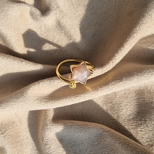 Könnte beinhalten: Ein goldfarbener Ring mit einem sternförmigen Edelstein. Der Stern hat einen hellrosa Farbton und ist in ein goldenes Band gefasst. Ein kleiner, strukturierter goldener Ball akzentuiert das Design des Rings. Der Ring wird auf einer beigen, strukturierten Oberfläche gezeigt.