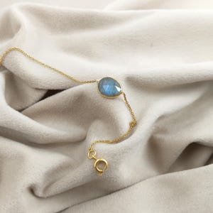 Op de afbeelding: Een delicate gouden kettingarmband met een peervormige, blauwe edelsteen. De edelsteen is gezet in een gouden bezel. De armband wordt getoond op een zachte, crèmekleurige stof.