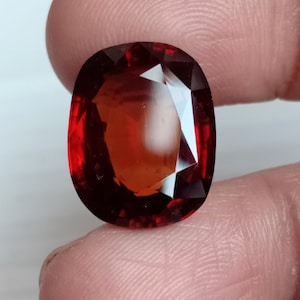 May include: A deep red gemstone with a faceted oval shape. The gemstone is held in a hand.