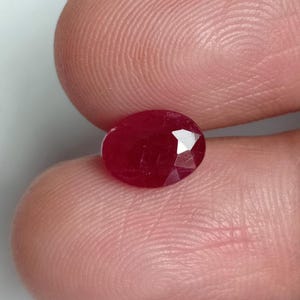 May include: A loose, oval-cut gemstone with a deep, saturated red color. The stone features a faceted surface that reflects light, showcasing a brilliant, polished finish. The gemstone is held between two fingers, highlighting its small size.