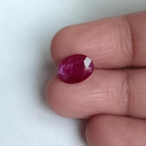May include: An oval-shaped, faceted ruby gemstone. The gem is a deep, rich red color with a polished surface. The ruby is held between fingers, showcasing its size and brilliance. This gemstone is suitable for jewelry.