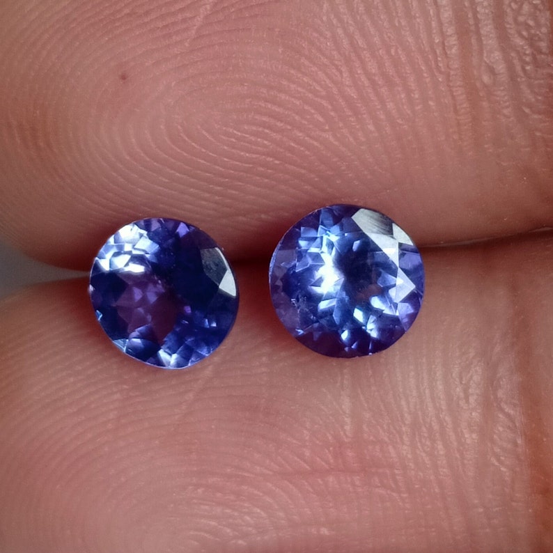 May include: Two round, faceted, purple gemstones with a blue hue. The gemstones are set in a bezel setting and are approximately 5mm in diameter.