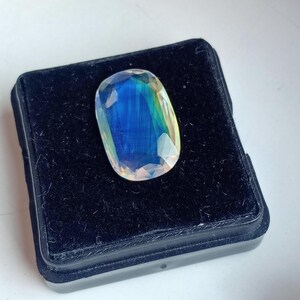 May include: A large, oval-shaped, iridescent gemstone with a blue, green, and yellow color scheme. The gemstone is displayed in a black velvet box.