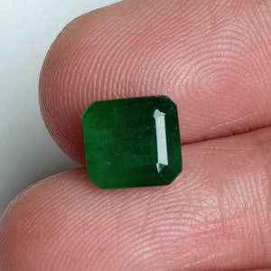 Puede incluir: Una gema de esmeralda verde vibrante, cortada en forma rectangular con bordes biselados. La gema está engastada en un engaste claro y facetado.