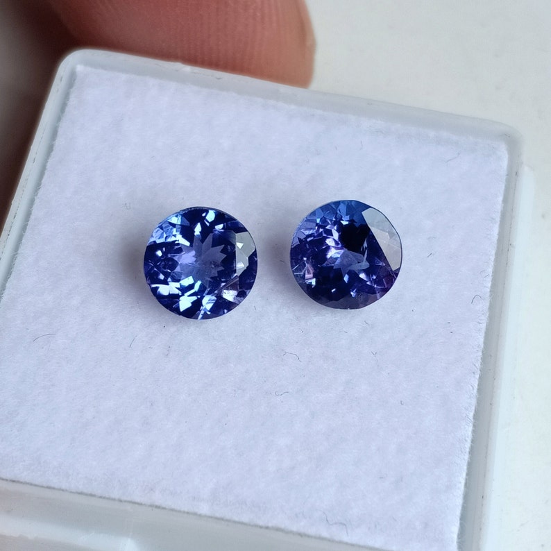 May include: Two round, blue gemstones with a purple hue, set in a white box. The gemstones are faceted and have a shiny finish.