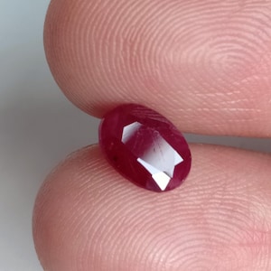 Puede incluir: Una piedra preciosa de color rojo intenso con talla ovalada y múltiples facetas que reflejan la luz. La piedra muestra un tono carmesí rico y saturado con una superficie pulida, destacando su claridad y su corte geométrico.