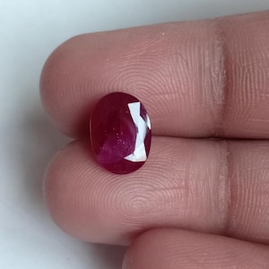 May include: A faceted, oval-shaped ruby gemstone. The ruby is a deep, rich red color with a polished surface. The gemstone is held between fingers, showcasing its size and brilliance.