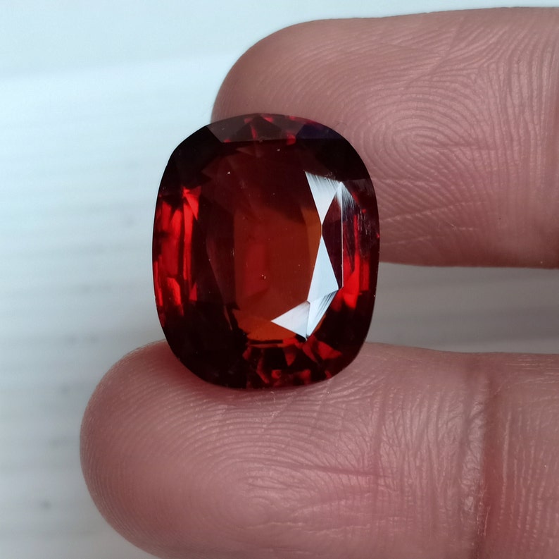 May include: A deep red gemstone with a cushion cut and a high polish. The gemstone is displayed on a finger.