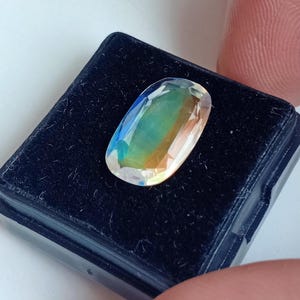 May include: A clear oval-shaped gemstone with iridescent colors, including green, blue, and yellow, displayed in a black velvet box.