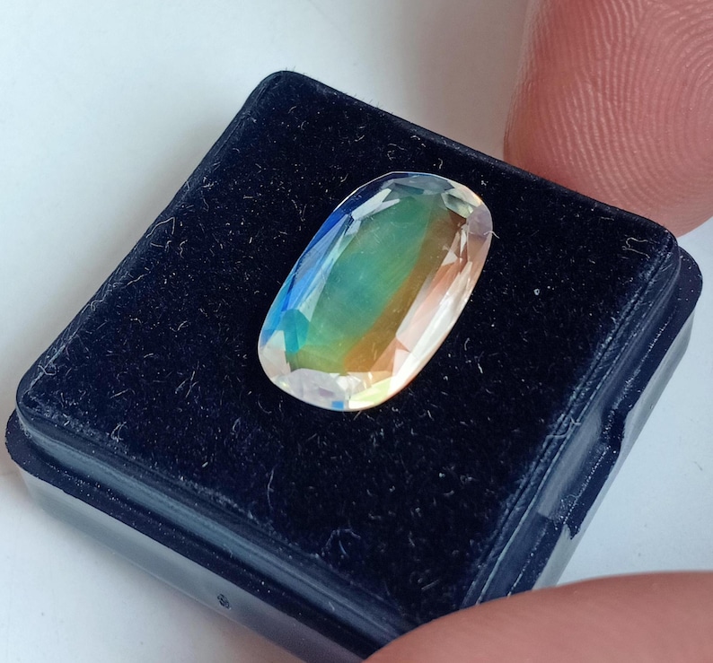 May include: A clear oval-shaped gemstone with iridescent colors, including green, blue, and yellow, displayed in a black velvet box.
