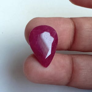 以下が含まれることがあります： ファセットカットされた、洋梨型のルビーの宝石。宝石は深く豊かな赤色で、光を捉える複数のファセットがあります。石は2本の指の間に挟まれ、そのサイズと形を示しています。この宝石は、自然なジュエリーの美しい例です。