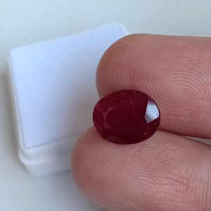 Pode incluir: Uma pedra preciosa de rubi facetada em forma oval. O rubi é de uma cor vermelha profunda e rica e é segurado entre dois dedos. Um objeto branco e retangular está no fundo.