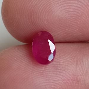 Può includere: Una gemma di rubino sfaccettata di forma ovale. Il rubino è di un colore rosso intenso e saturo con molteplici sfaccettature che riflettono la luce. La gemma è tenuta tra due dita.