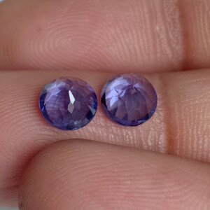 May include: Two round, faceted, purple gemstones. The gemstones are approximately the same size and have a light purple hue.