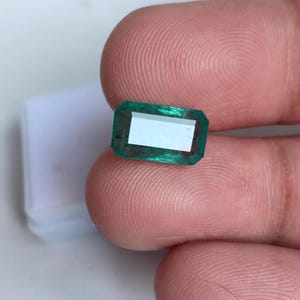 May include: A vibrant green emerald gemstone, cut in a rectangular shape with bevelled edges. The gemstone is held in a hand, showcasing its clarity and brilliance.