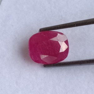 May include: A faceted, cushion-cut ruby gemstone, held by tweezers. The ruby is a deep, saturated red color, with visible internal inclusions and a polished surface. The gemstone is set against a white background.