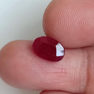 Pode incluir: Uma pedra preciosa facetada de formato oval com uma cor vermelha profunda e saturada. A pedra apresenta uma superfície polida com múltiplas facetas geométricas que refletem a luz, exibindo uma qualidade translúcida e um tom carmesim vibrante e rico.