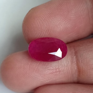 Puede incluir: Una gema de corte ovalado vibrante con un tono rojo magenta profundo y saturado. La piedra presenta una superficie facetada que refleja la luz, creando reflejos brillantes distintos en su estructura cristalina pulida, translúcida y suave.