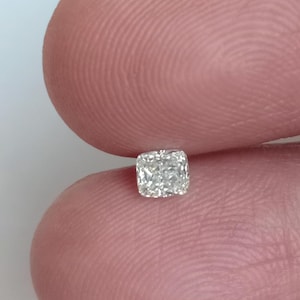 Könnte beinhalten: Nahaufnahme eines kleinen, quadratisch geschliffenen Diamanten mit einer brillanten, facettenreichen Oberfläche. Der Diamant wird zwischen zwei Fingern gehalten, was seine Klarheit und sein Funkeln zeigt.