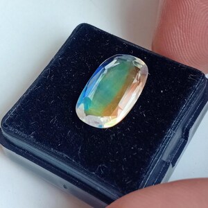May include: A clear oval-shaped gemstone with iridescent colors, including blue, green, and yellow, displayed in a black velvet box.