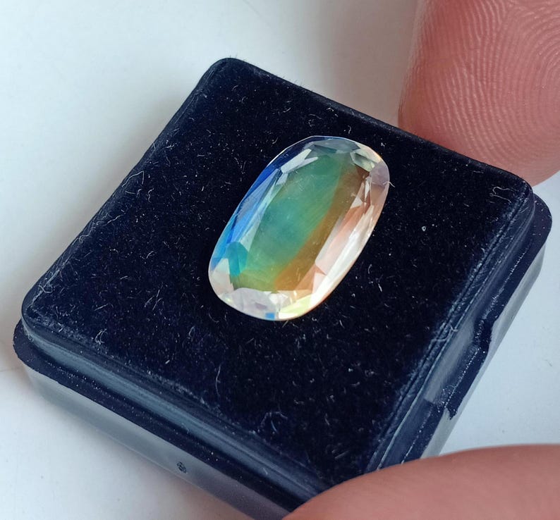 May include: A clear oval-shaped gemstone with iridescent colors, including blue, green, and yellow, displayed in a black velvet box.