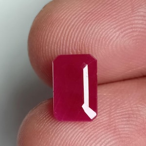 May include: A vibrant red gemstone with a rectangular emerald cut. The gemstone is set in a clear, faceted setting.