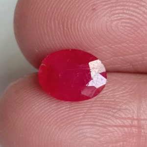 Puede incluir: Una gema ovalada de color rojo vibrante con un corte facetado. La gema está colocada sobre un fondo rosa.